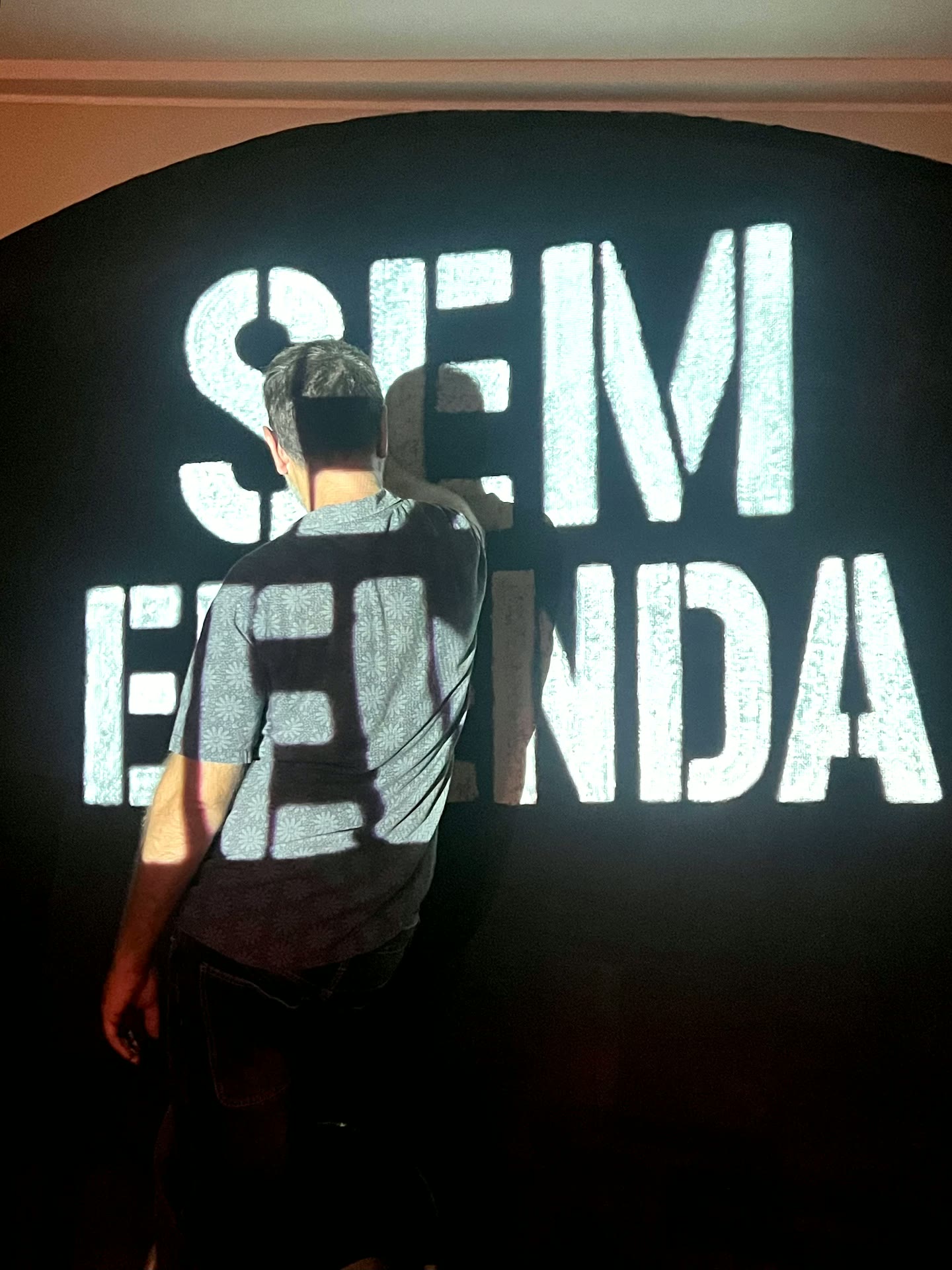 Pessoa de costas diante do lettering Sem Emenda projetado numa parede escura