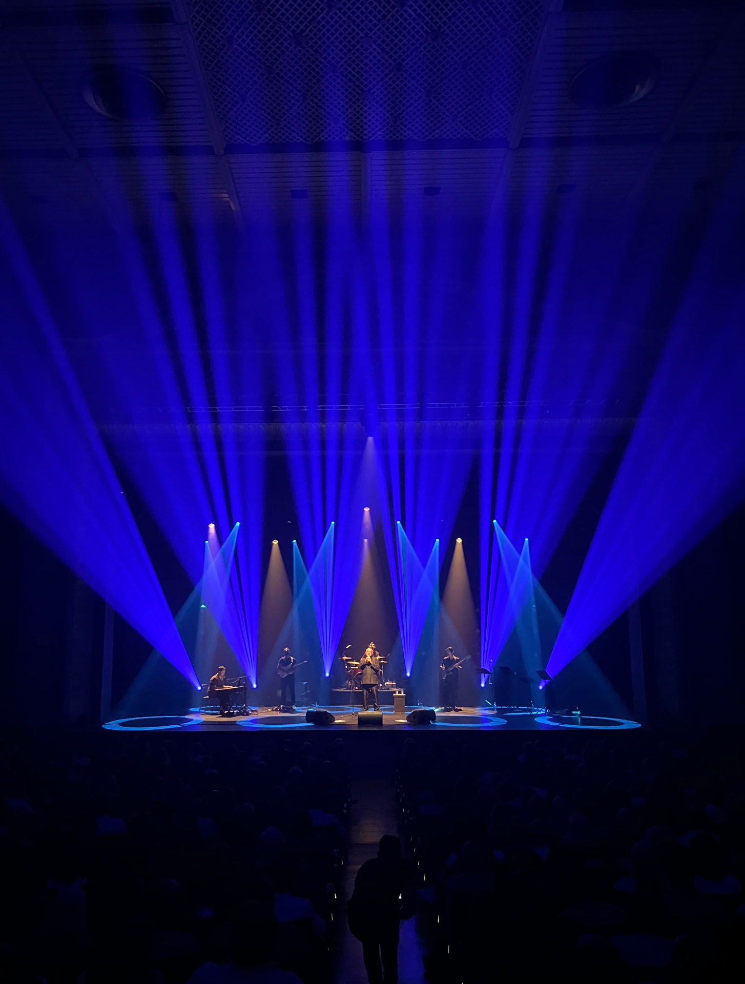 Sala de espetáculos com feixes de luz sobre o palco e uma figura no corredor central